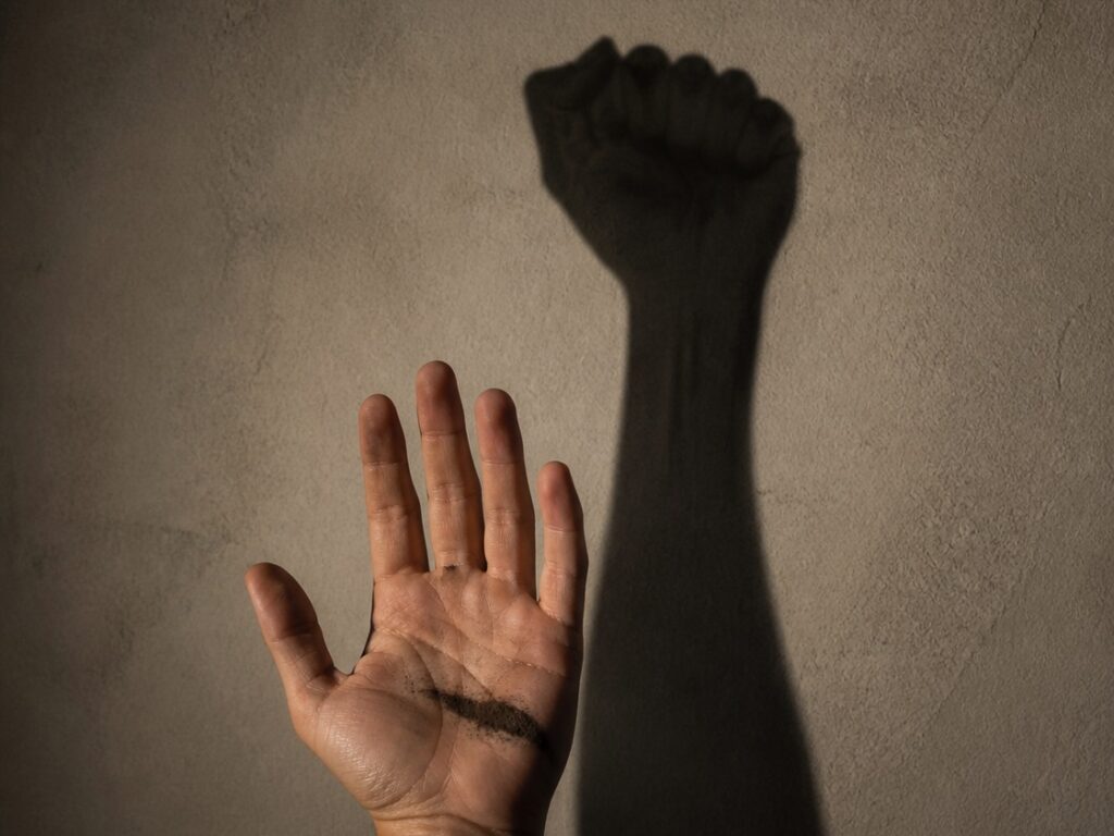 Eine erhobene offene Hand vor einer hellen Wand; ihr Schatten an der Wand zeigt eine geballte Faust.