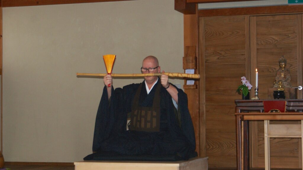 Der Shuso hält bei der Dharma-Befragung den Stab mit dem Fächer, dahinter der Altar mit Kerze und Buddhafigur.