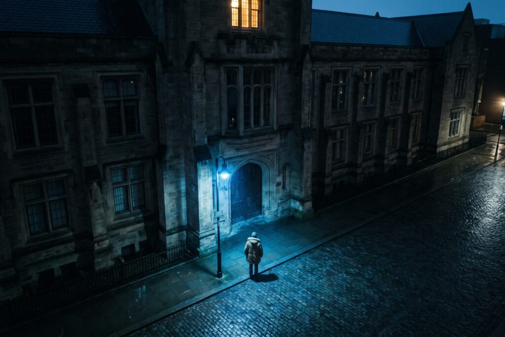 Nachts steht eine einzelne Person im Mantel unter einer blauen Straßenlaterne auf dem nassen Pflaster vor dem Tor eines dunklen Universitätsgebäudes, hoch über ihr leuchtet ein einziges Fenster warm.