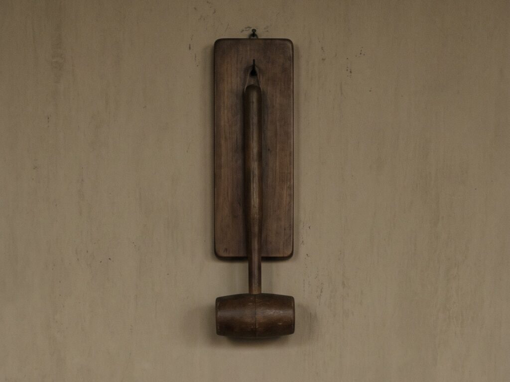 An einer hellen, fleckigen Putzwand hängt ein hölzerner Schlägel an einem schmalen Brett, der schwere Kopf nach unten. Das Holz ist dunkel und abgegriffen, die Wand ringsum bleibt leer.